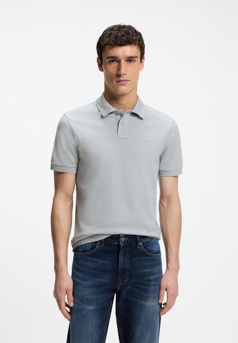 Homme portant un polo gris clair et un jean bleu foncé, debout devant un fond blanc, regardant droit devant avec une expression neutre.