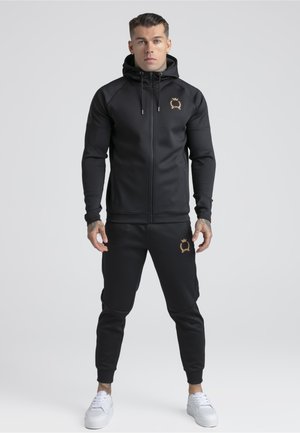 Mann som står iført svart hettegenser med glidelås og matchende joggebukser med gullfargede våpenskjoldlogoer, kombinert med hvite sneakers, på ensfarget lys bakgrunn.