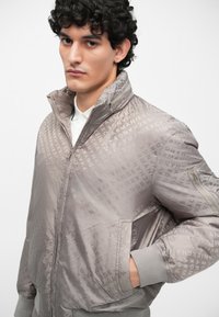 Jachetă bomber gri cu model logo embosat, guler înalt, fermoar frontal și manșete ribbate. Material neted, ușor, cu o croială structurată.
