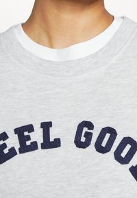 Gros plan sur une personne portant un sweat-shirt gris clair par-dessus une chemise blanche, avec le texte "FEEL GOOD" en lettres bleu marine sur la poitrine.