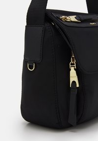 Černá crossbody taška se zlatým zipem se značkou "DKNY", přední kapsou pod klopou a nastavitelním popruhem se zlatým kovovým detailem.