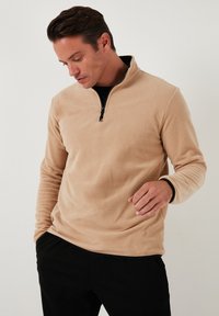 Beež villane pullover musta tõmbluku kaelusega ja tekstuurse pinnaga. Pikad varrukad kontrastsete mansettide detailiga. Kantud musta särgi peal.