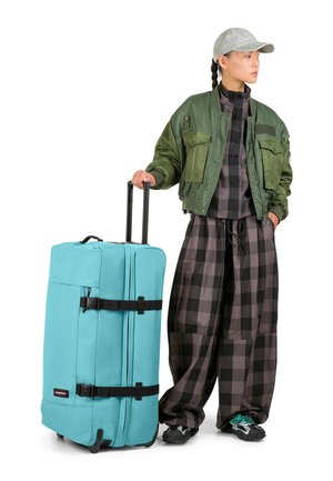 Grande valigia turchese con cinghie nere, ruote e manico. L'utente indossa una giacca bomber verde oliva e pantaloni a quadri neri.