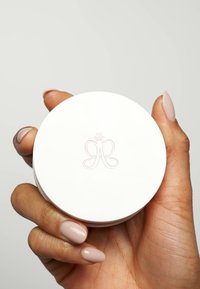 Witte ronde container vastgehouden in een hand, met een verhoogd vlinderlogo en een gladde matte afwerking. Geen zichtbare patronen of versieringen.