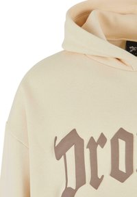 Beige huppari, jossa on suuri ruskea graafinen teksti etupuolella, pehmeällä pinnalla ja klassisella kengurutaskustyylillä.