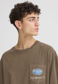 T-shirt oversize en coton marron avec un col rond, doté d'une poche avant et d'un texte brodé : "SoulScape - Hors du bureau depuis 99."