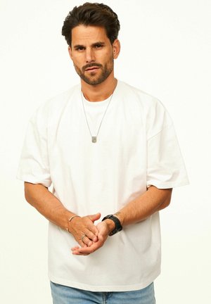 Man with dark hair and beard wearing a white oversized t-shirt, silver necklace, wristwatch, and light blue jeans standing against a plain background.