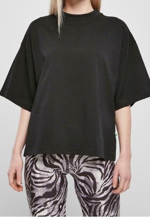 Persoon draagt een loszittend zwart T-shirt en zebra gestreepte leggings, staand met ontspannen armen langs het lichaam tegen een effen achtergrond.