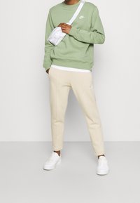 Ljusgrön sweatshirt med vitt Nike-logotyp, beige tapered byxor med logotyp, och vita sneakers. Axelremsväska i vitt. Mjukt, texturerat tyg.