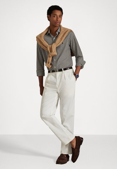 Pantalons blancs, chemise à carreaux boutonnée, et un pull en tricot beige drapé sur les épaules. Des mocassins marron complètent la tenue.