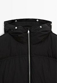 Schwarze gepolsterte Jacke mit Kapuze, ausgestattet mit silbernen Beschlägen, einem Frontreißverschluss und sichtbaren Nähdetails. Glatte Textur, leichtes Design.
