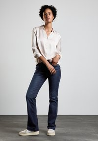 Blouse en satin rose clair avec bouton sur le devant, associée à un jean bleu foncé et des baskets beiges. Texture lisse et coupe décontractée, avec des manches retroussées.