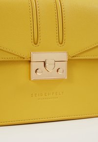 Gelbe Lederhandtasche mit einem goldfarbenen Hardwareverschluss, strukturiertem Oberflächenfinish und geprägtem Markenzeichen am Boden.