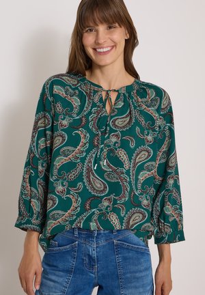 Femme souriante portant un chemisier vert à motif cachemire avec un col à nouer et un jean en denim bleu sur un fond clair uni.