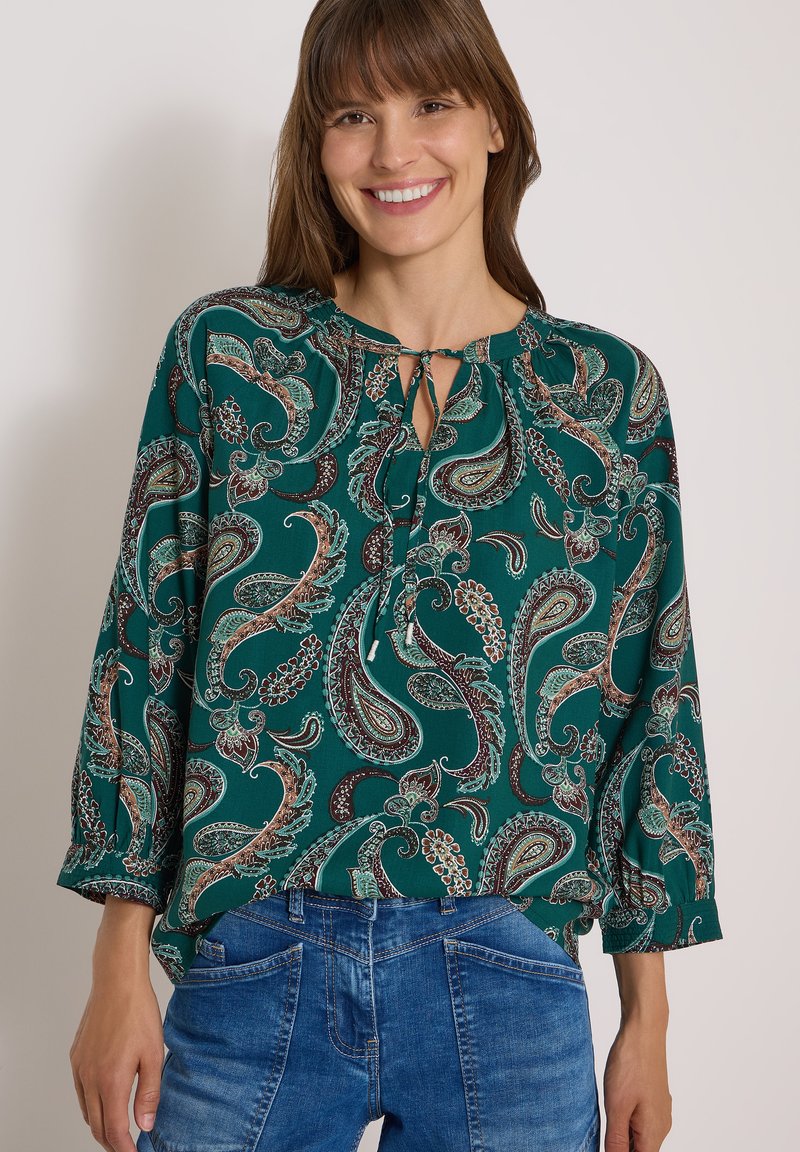 Femme souriante portant un chemisier vert à motif cachemire avec un col à nouer et un jean en denim bleu sur un fond clair uni.