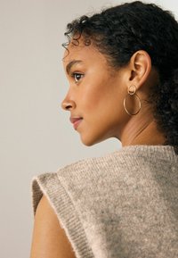 Kuldsete ringide kõrvarõngad, millel on ümmargune disain, kaunistavad mudelit, kellel on sile ja läikiv viimistlus. Mudel kannab heledat kudumist kampsunit.