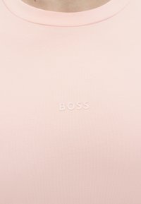 Κοντινό πλάνο ενός ανοιχτό ροζ τοπ με τη λέξη «BOSS» εγχαραγμένη στο στήθος κάτω από στρογγυλή λαιμόκοψη.