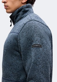 Homme portant une veste zippée bleu-gris texturée avec un col montant et un petit patch noir sur le haut de la manche.