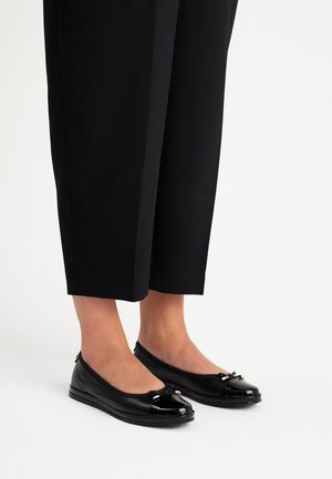 Czarne baletki z błyszczącym czubkiem i ozdobną kokardą, wykonane z gładkiej skóry, w połączeniu z czarnymi, skróconymi spodniami.