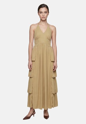 Femme debout face à l'avant, portant une robe longue beige sans manches à col halter avec des poches latérales en étages et des talons pointus marron.