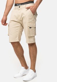 Shorts cargo beiges avec une fermeture à bouton et à zip, poches latérales et une finition texturée. Portés avec des baskets blanches et une ceinture de marque noire.