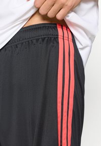 Μαύρο αθλητικό σορτς από λείο ύφασμα, με τρεις κατακόρυφ ες κόκκινες ρίγες κατά μήκος της πλευράς και ελαστική μέση.