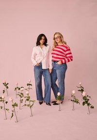 Zwei Frauen stehen vor einem pinkfarbenen Hintergrund, umgeben von Vasen mit rosa Rosen, sie tragen legere Jeans und langärmlige Oberteile, eines gestreift und das andere einfarbig.