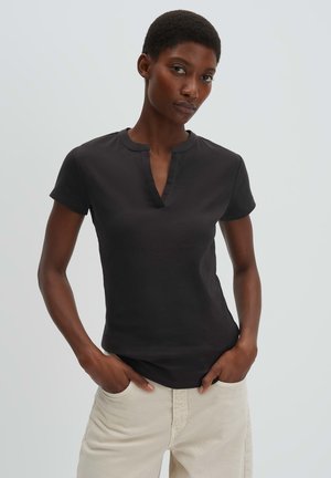 Frau mit kurzen Haaren, die ein schwarzes Kurzarmoberteil mit einem Ausschnitt und hellbeige Hose trägt, stehend mit den Händen in den Taschen.