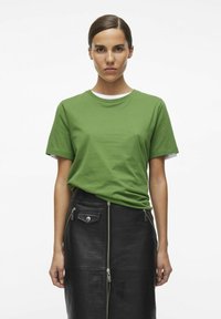 Camiseta de algodón verde superpuesta sobre una blusa blanca, combinada con una falda de cuero negra con cierres de cremallera y un acabado texturizado.