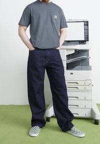 Szara bawełniana koszulka z kieszenią na piersi, granatowe dżinsy o szerokich nogawkach oraz zielone, w kratę buty wsuwane. W tle drukarka biurowa.