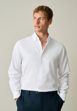 Uomo con camicia bianca abbottonata e pantaloni scuri, che guarda leggermente di lato con le mani in tasca su uno sfondo semplice.