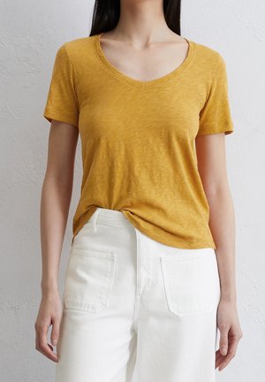 Musztardowa krótko rękawowa koszulka z dekoltem w serek i teksturowanym materiałem, zestawiona z białymi spodniami o wysokim stanie z tylnymi kieszeniami.