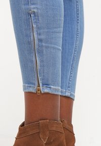 Pantalones de mezclilla azul claro cortos con detalles de cremallera lateral y un diseño ajustado. Combinados con botas de ante marrones hasta el tobillo.