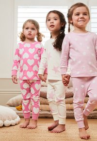 Trois enfants en pyjamas pastel : un en rose avec des cœurs, un en blanc avec des étoiles roses, et un en rose avec des pois blancs. Texture douce en coton.
