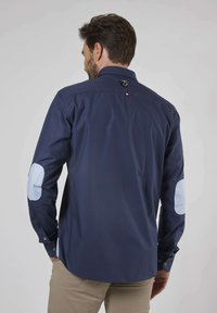 Chemise bleu marine avec un col classique, des manches longues et des coudières contrastantes bleu clair. Elle dispose de poignets à boutons et d'un patch logo.