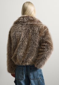 Chaqueta de pelaje sintético en marrón claro con una textura suave y un cuello ancho. Presenta un corte corto y mangas rectas. Se lleva con pantalones de denim.