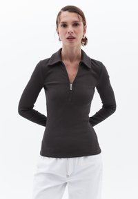 Langärmliges Oberteil aus dunkelbraunem Rippstoff, mit einem Reißverschluss-Halsausschnitt und einem kragenartigen Design. Kombiniert mit weißen Hosen.