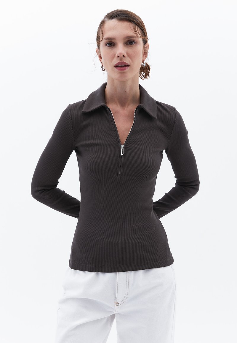 Langärmliges Oberteil aus dunkelbraunem Rippstoff, mit einem Reißverschluss-Halsausschnitt und einem kragenartigen Design. Kombiniert mit weißen Hosen.