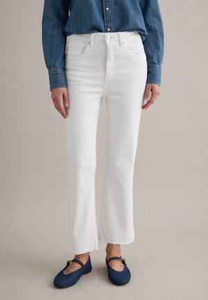 Persona che indossa jeans bianchi a gamba dritta, camicia di jeans blu e scarpe Mary Jane blu navy su sfondo neutro.