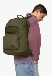 Zaino verde scuro con molte tasche con cerniera, dettagli in rete e spallacci regolabili. Indossato sulla schiena, abbinato a una maglietta di colore viola chiaro.