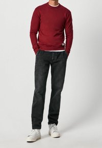 Röd långärmad tröja med rund halsringning, bärs över en vit skjorta. Utanpå svarta jeans och vita sneakers. Enkel design, minimala detaljer.