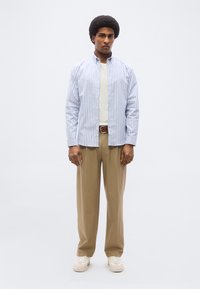 Άνδρας με αφρο μαλλί που φοράει μπεζ παντελόνια, λευκά αθλητικά παπούτσια, λευκό μπλουζάκι και ανοιχτό πουκάμισο με μπλε και λευκές ρίγες.