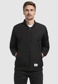 Schwarze Bomberjacke mit Reißverschluss vorne, geripptem Kragen und zwei Seitentaschen. Mit strukturierten Akzenten an den Ärmeln und einem Logopatch am Saum.