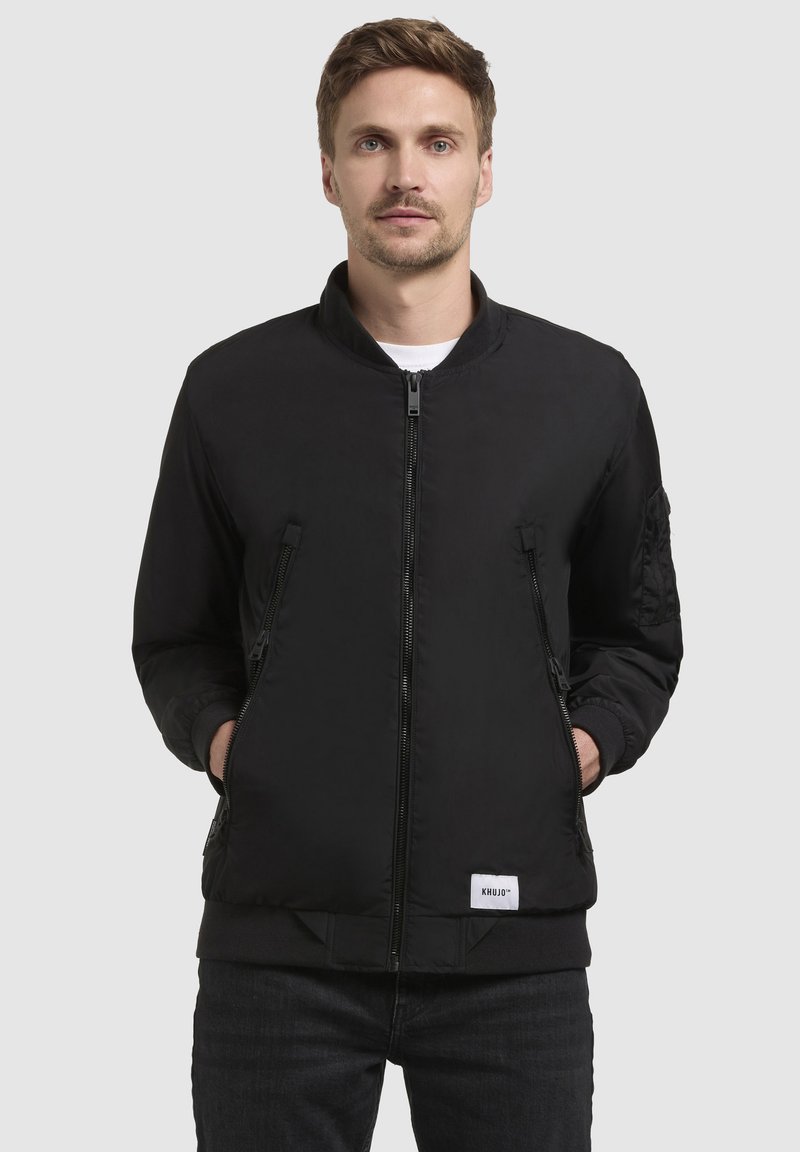 Schwarze Bomberjacke mit Reißverschluss vorne, geripptem Kragen und zwei Seitentaschen. Mit strukturierten Akzenten an den Ärmeln und einem Logopatch am Saum.