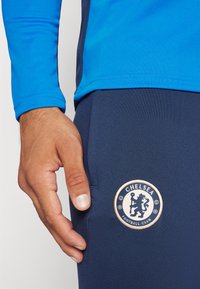 Niebieska bluzka z długim rękawem połączona z granatowymi spodniami, z okrągłym logo Chelsea Football Club przedstawiającym lwa i piłkę.