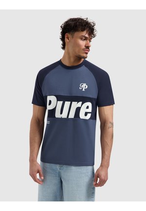 Man met krullend haar draagt een marineblauw en blauw T-shirt met "Pure" in grote witte letters, gecombineerd met lichtblauwe spijkerbroek, kijkt opzij.