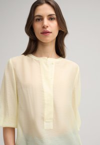 Blusa giallo chiaro con scollatura rotonda, maniche a tre quarti e una placchetta con bottoni. Il tessuto ha una finitura morbida e leggermente strutturata.