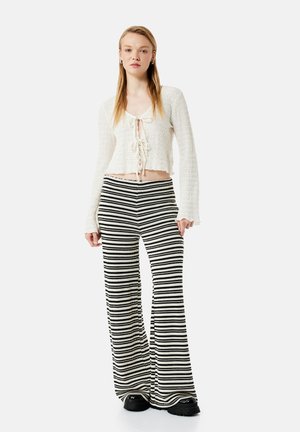 Młoda kobieta stoi przodem, ubrana w biały teksturowany top z długim rękawem i wiązaniem z przodu oraz czarno-białe szerokie spodnie w paski.