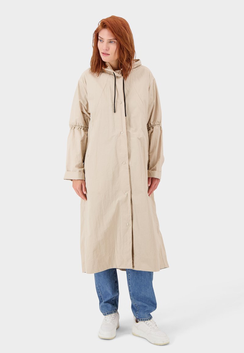 Beige Kapuzenmantel aus leichtem Stoff mit elastischen Bündchen, Knopfverschluss und Kordelzugdetails. Getragen über blauen Jeans und weißen Sneakers.