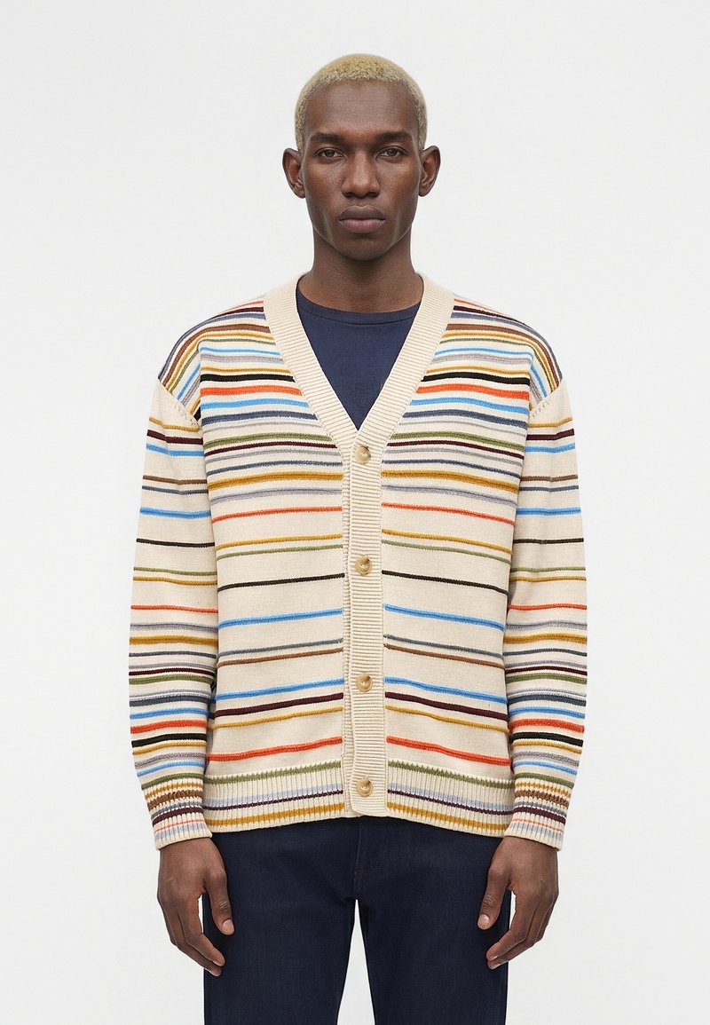 Νέος άνδρας με κοντά ξανθά μαλλιά, φοράει μια κρεμ ζακέτα με πολύχρωμες οριζόντιες ρίγες και σκοτεινό παντελόνι, στέκεται μπροστά σε λευκό φόντο.